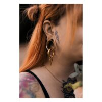 Femme aux cheveux rouges portant un Labret Push-Fit Monstera doré à l'oreille, tatouages visibles