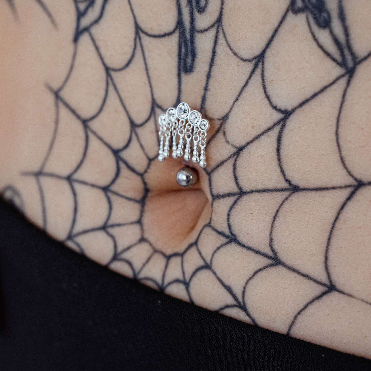 Micro Bananabell en argent avec cristaux Piercing Bananabell en argent avec cinq cristaux suspendus dans un nombril, entouré d'un tatouage toile d'araignée