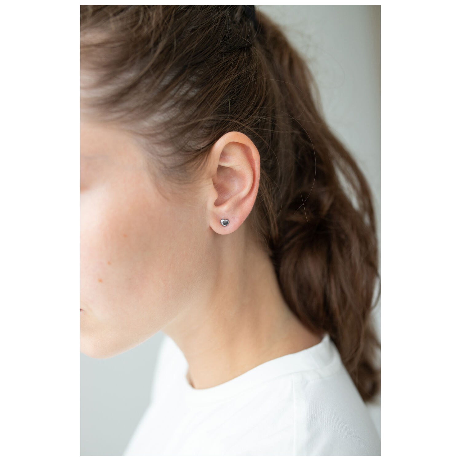Boucles d'oreilles cœur argentées sur l'oreille d'une femme aux cheveux bruns, fond blanc