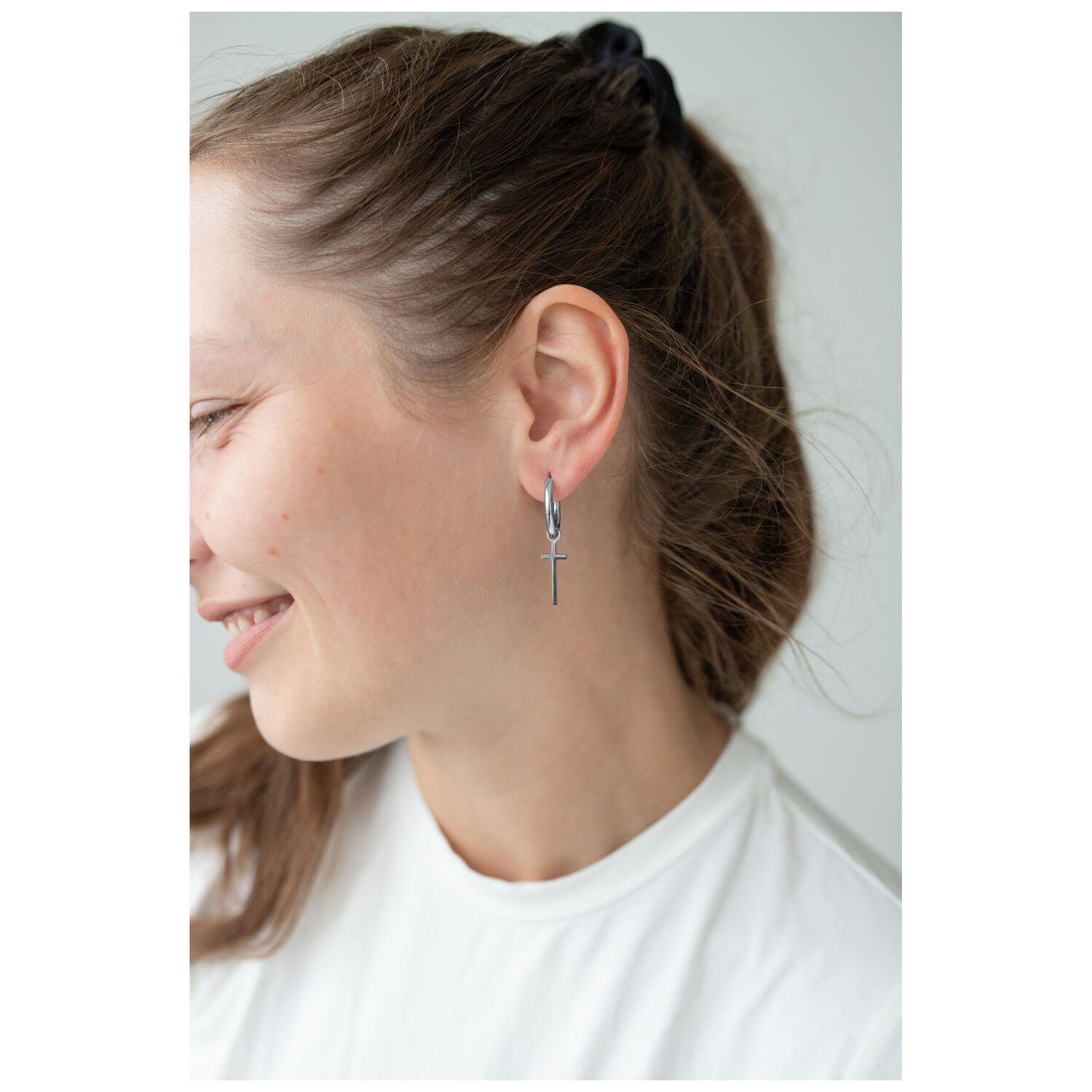 Boucles d'oreilles Little Cross élégantes en argent Femme portant des boucles d'oreilles en croix argentées, souriante de profil sur fond clair