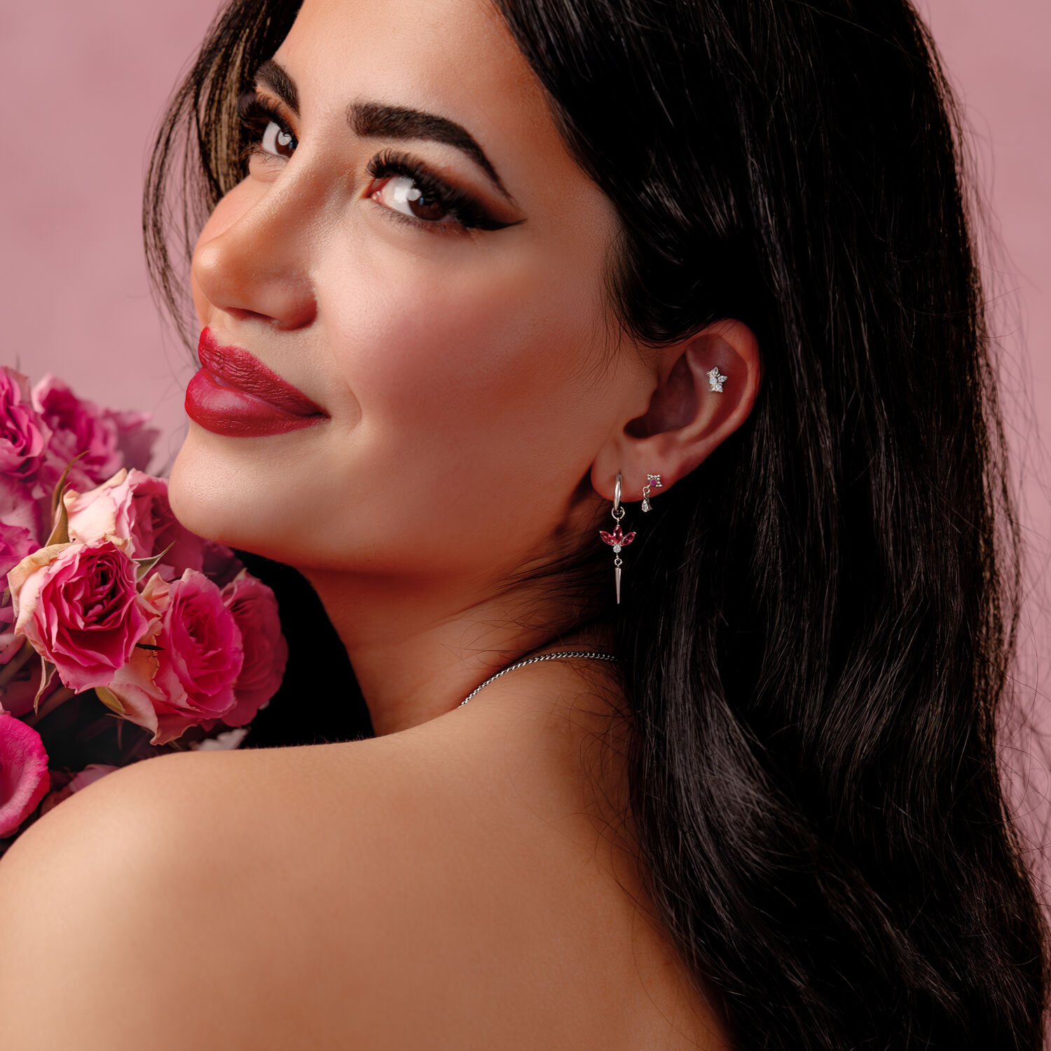Femme aux longs cheveux bruns portant des boucles d'oreilles argentées avec des cristaux rouges, tenant des roses roses devant un fond rose.