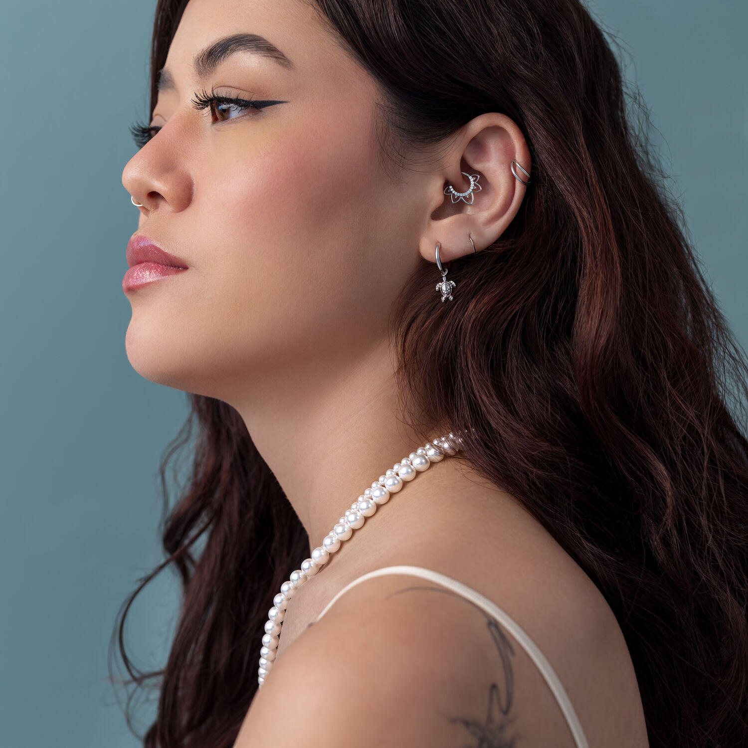 Boucles d'oreilles tortue argentées avec collier de perles Femme aux longs cheveux bruns, boucles d'oreilles tortue argentées, collier de perles, fond bleu