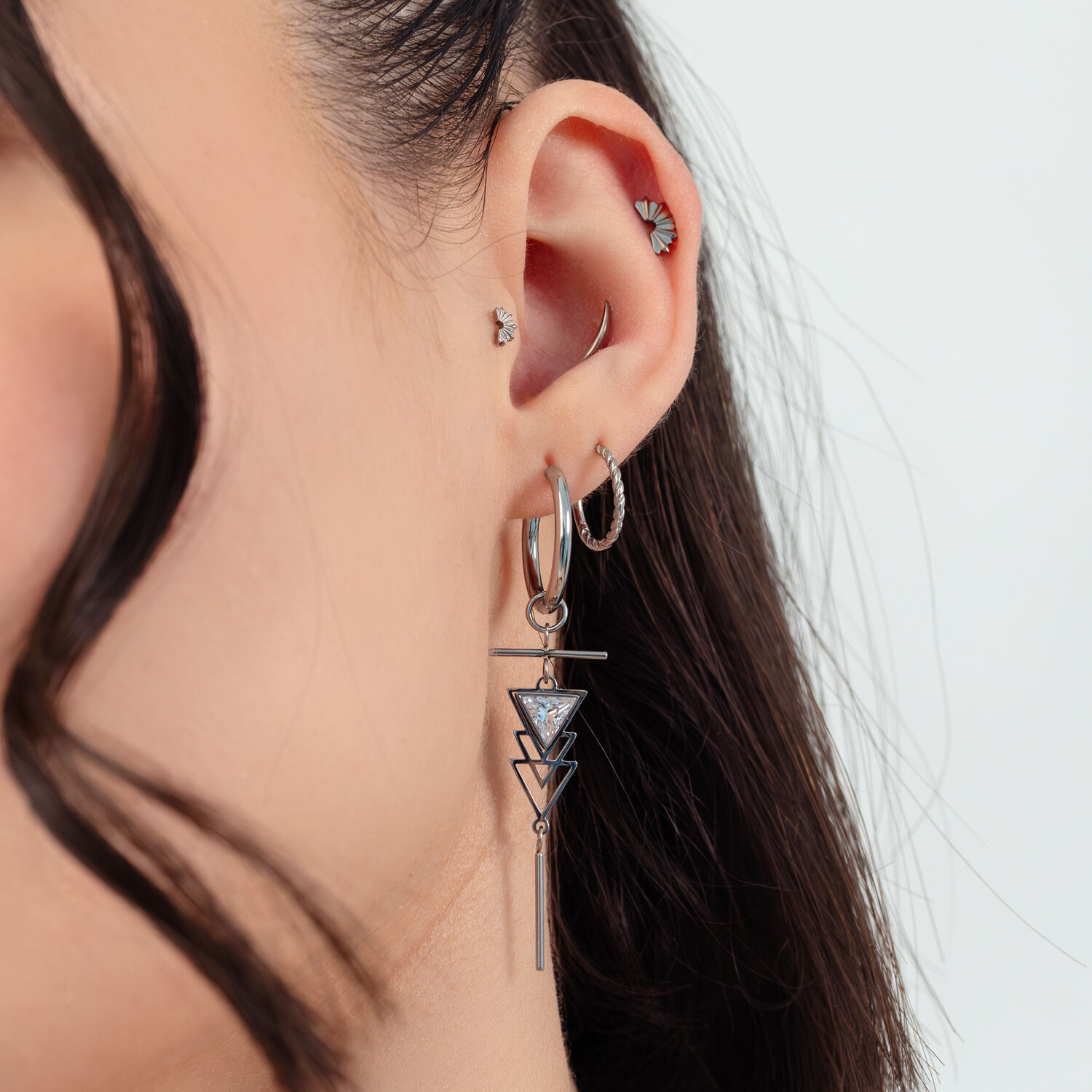 Boucles d'oreilles Art déco argentées avec détails pendants Femme avec des boucles d'oreilles Art déco argentées et plusieurs piercings devant un fond blanc