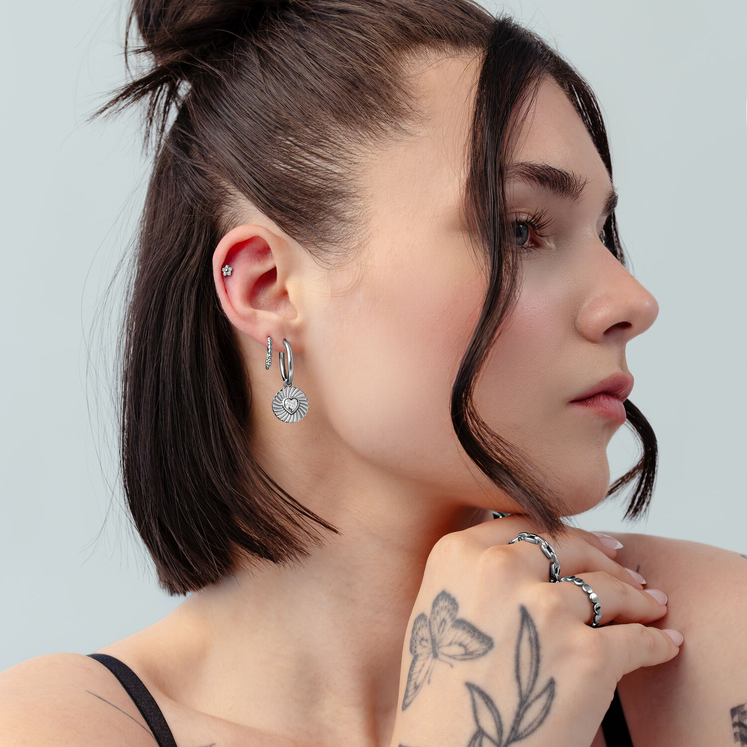 Créoles Cœur Argentées Riffled en Profil Femme aux cheveux courts et foncés portant des créoles argentées avec design cœur et tatouages sur fond gris clair.