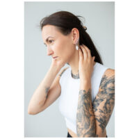 Petites boucles d'oreilles Hamsa portées à l'oreille Femme tatouée portant des boucles d'oreilles Hamsa, top blanc, fond gris