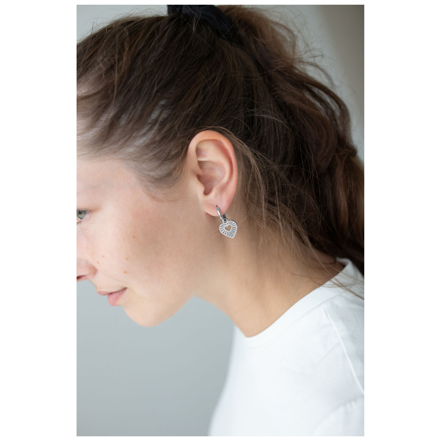 Petites créoles cœur en acier chirurgical Femme portant des créoles cœur argentées, photographiée de côté, sur fond blanc