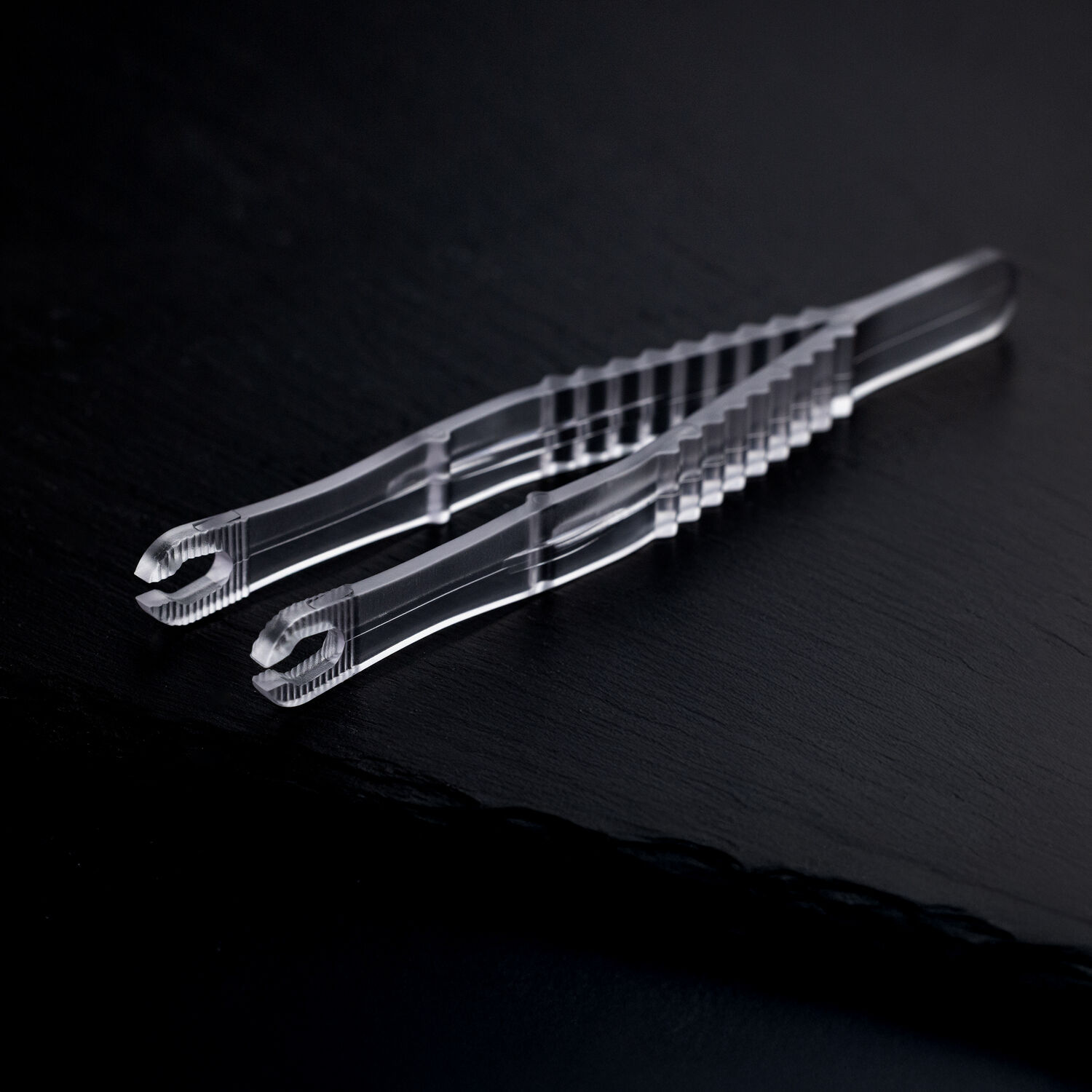 Deux pinces à piercing rondes et stériles transparentes croisées sur un fond sombre.