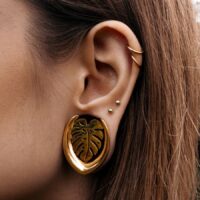 Boucles d'oreilles Monstera dorées, portées à l'oreille, avec des piercings en arrière-plan