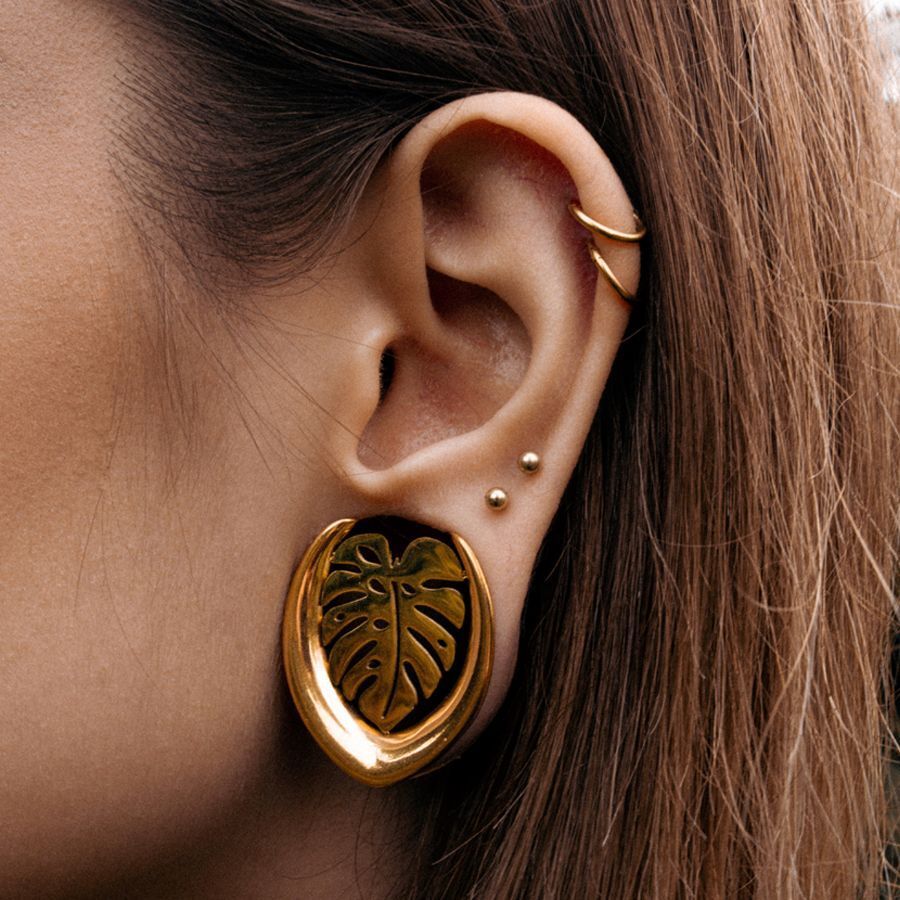 Boucles d'oreilles Monstera dorées, portées à l'oreille, avec des piercings en arrière-plan
