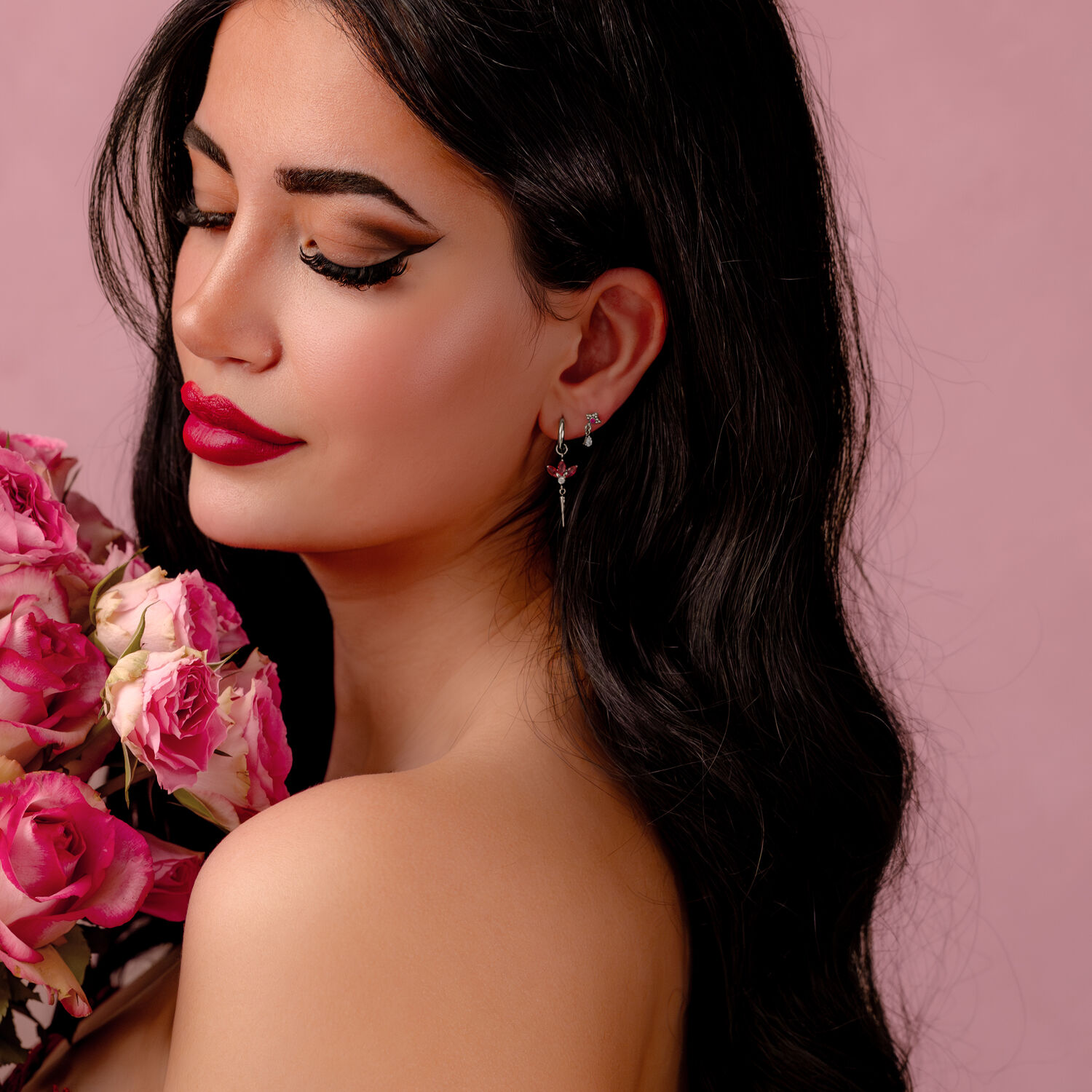 Boucles d'oreilles argentées avec cristaux rouges Femme aux longs cheveux noirs avec des boucles d'oreilles argentées et des cristaux rouges, tenant des fleurs roses sur fond rose