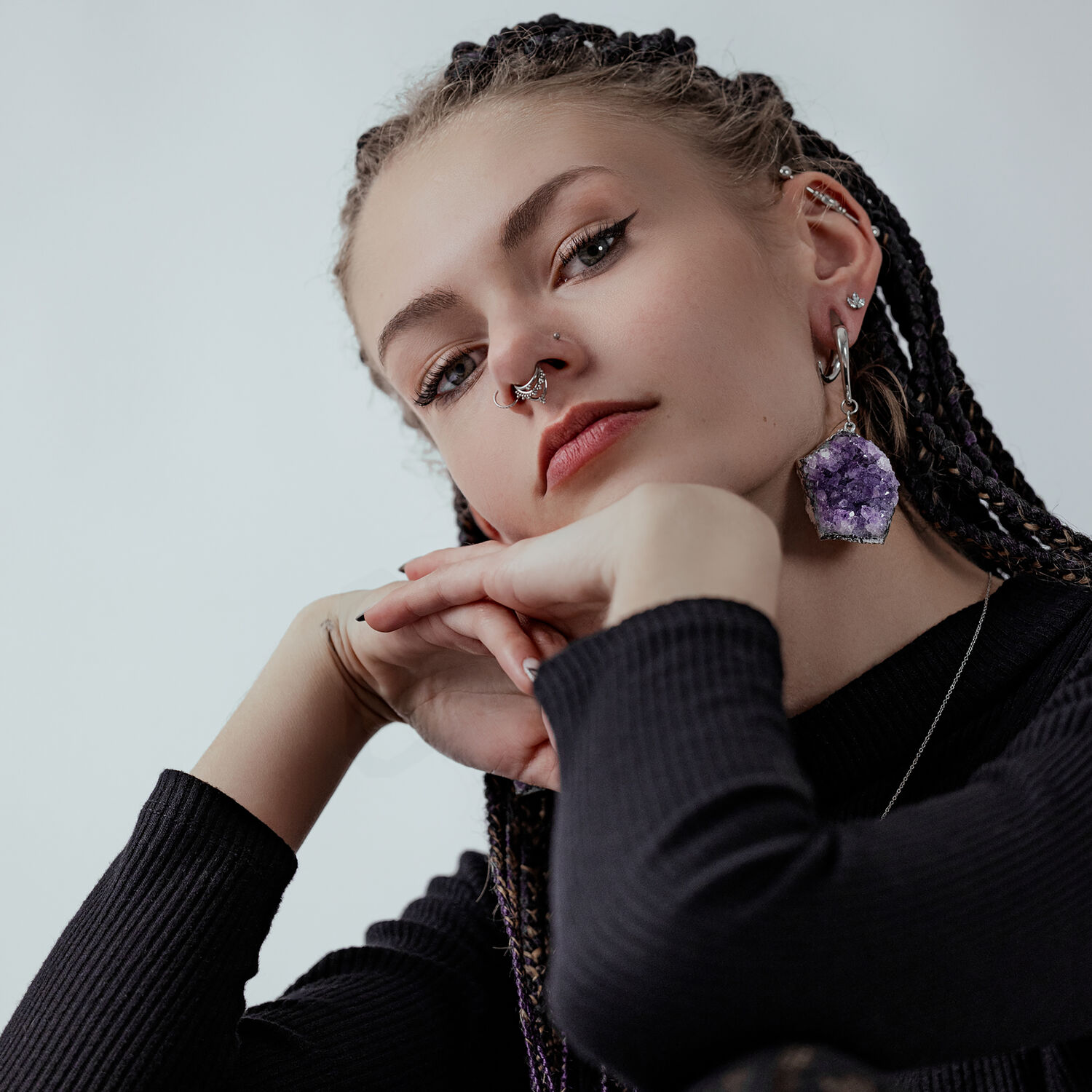 Femme avec boucle d'oreille en améthyste et piercing au nez Jeune femme avec des tresses, boucle d'oreille en améthyste, piercing au nez, pull noir sur fond gris