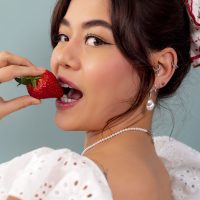 Femme avec fraise et piercings Jeune femme tenant une fraise à la main, plusieurs boucles d'oreilles et un collier, sur fond bleu