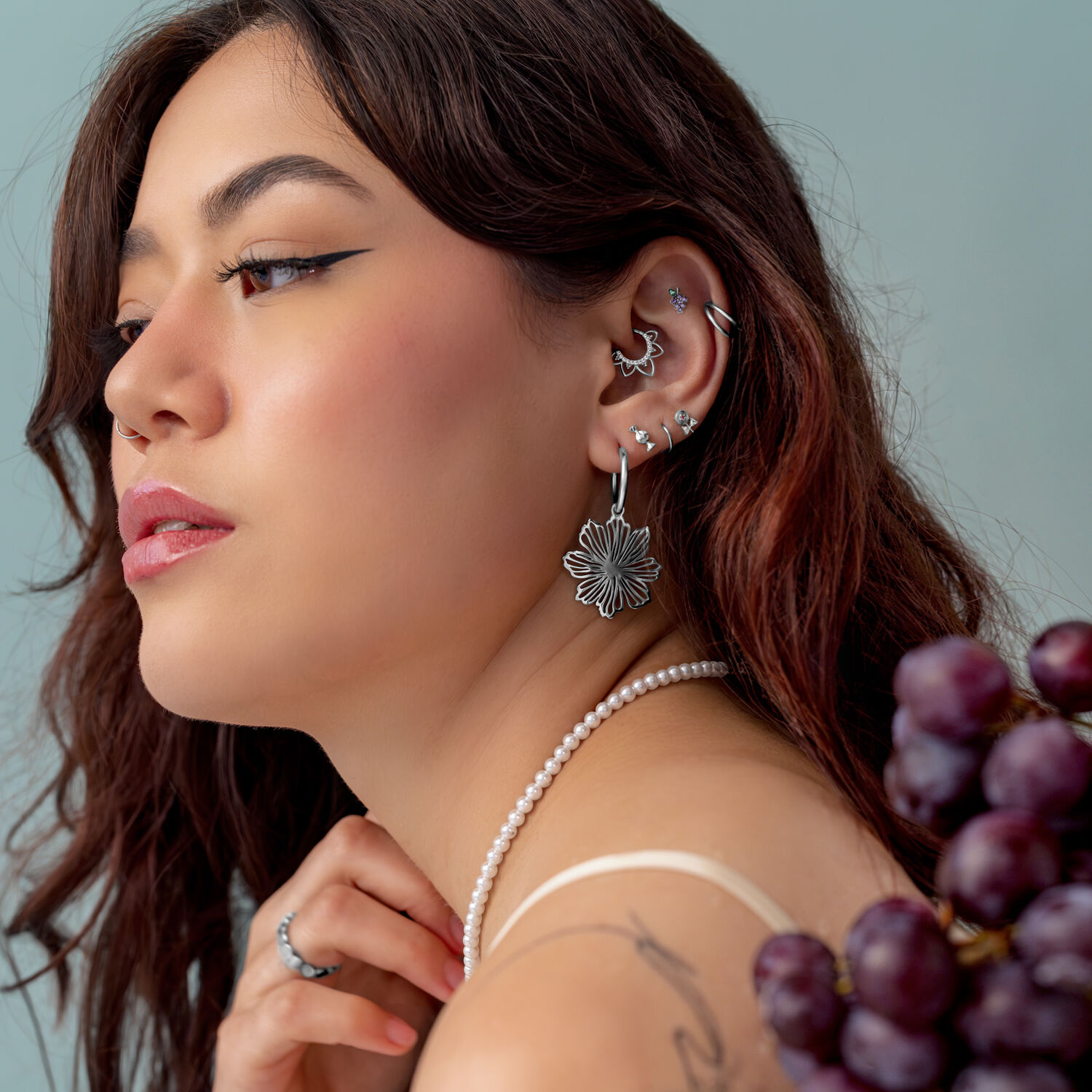 Embout Grape Push Fit Argenté Femme avec plusieurs piercings en argent à l'oreille, portant un collier de perles et tenant des raisins.