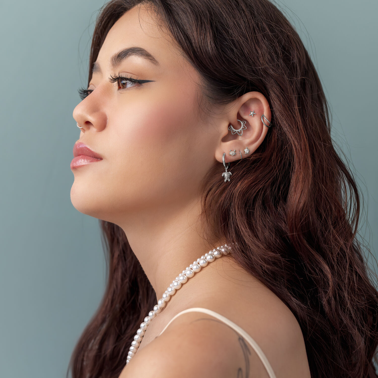 Embout Tortue Push Fit Argenté pour l'Oreille Femme aux cheveux bruns portant plusieurs piercings en argent et un pendentif en perles sur fond bleu