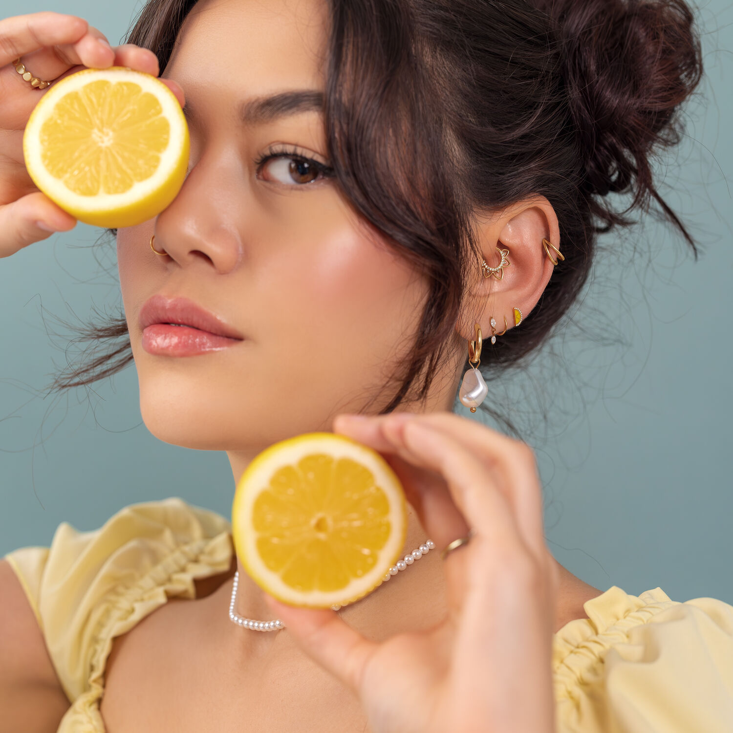 Femme avec des citrons et des piercings d'oreille Femme tenant un demi-citron devant son œil, portant des piercings d'oreille en or, sur fond bleu