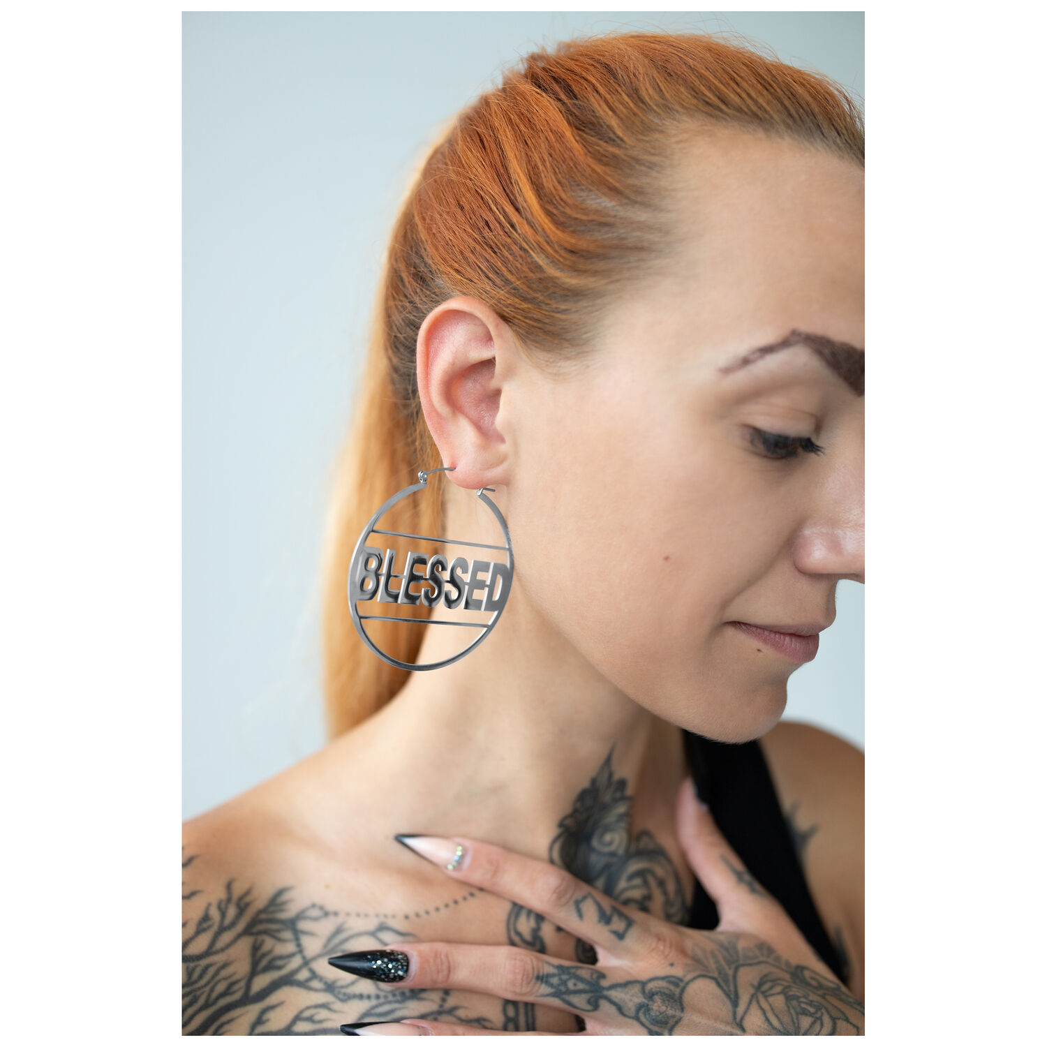 Boucles d'oreilles Blessed Hoops argentées portées à l'oreille Boucles d'oreilles Blessed Hoops argentées en acier inoxydable, portées à l'oreille d'une femme, sur fond neutre