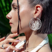 Noble Leaf Hoops en argent à l'oreille Boucles d'oreilles Noble Leaf Hoops en argent sur une femme avec des cheveux tressés et un fond tropical