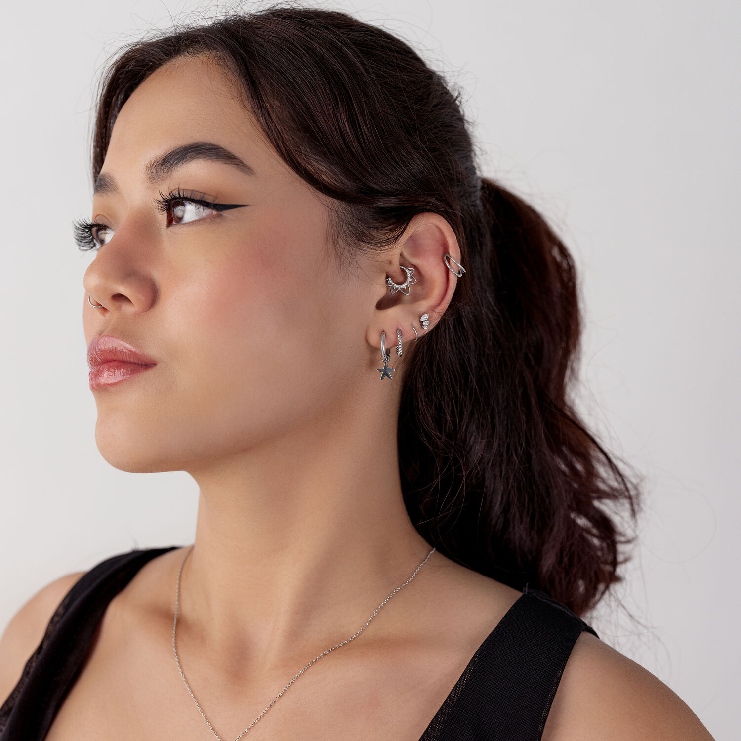Femme aux cheveux noirs portant un clicker de septum noir et plusieurs boucles d'oreilles de différentes formes sur un fond blanc.