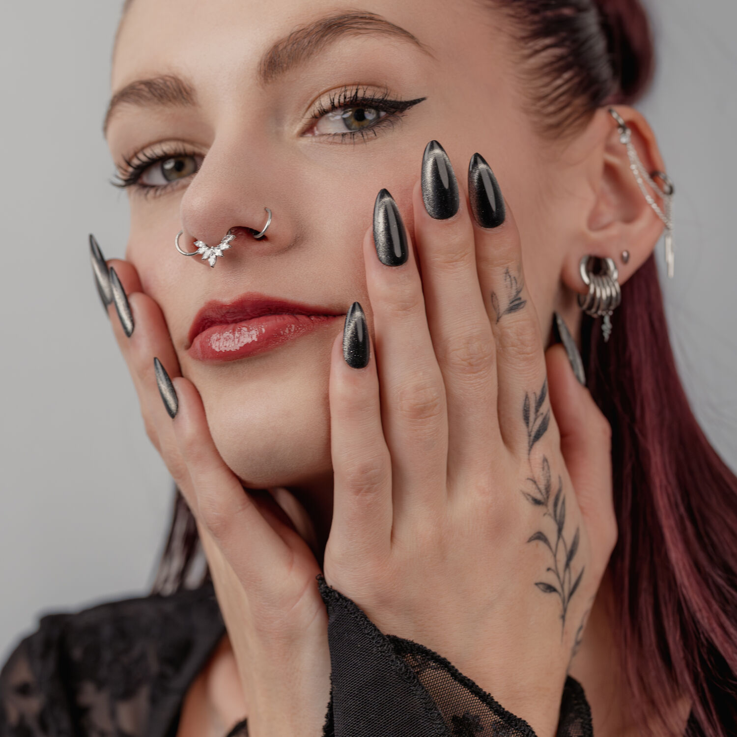 Design d'ongles sombre et piercings Femme avec des ongles noirs et brillants, plusieurs piercings aux oreilles et au nez, sur fond gris