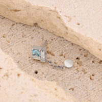 Clou d'oreille argenté avec pendentif chaîne Clou d'oreille argenté avec pierre carrée cristalline et pendentif chaîne sur pierre beige.
