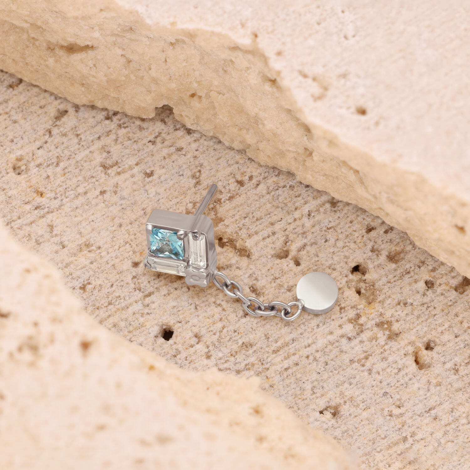 Clou d'oreille argenté avec pendentif chaîne Clou d'oreille argenté avec pierre carrée cristalline et pendentif chaîne sur pierre beige.