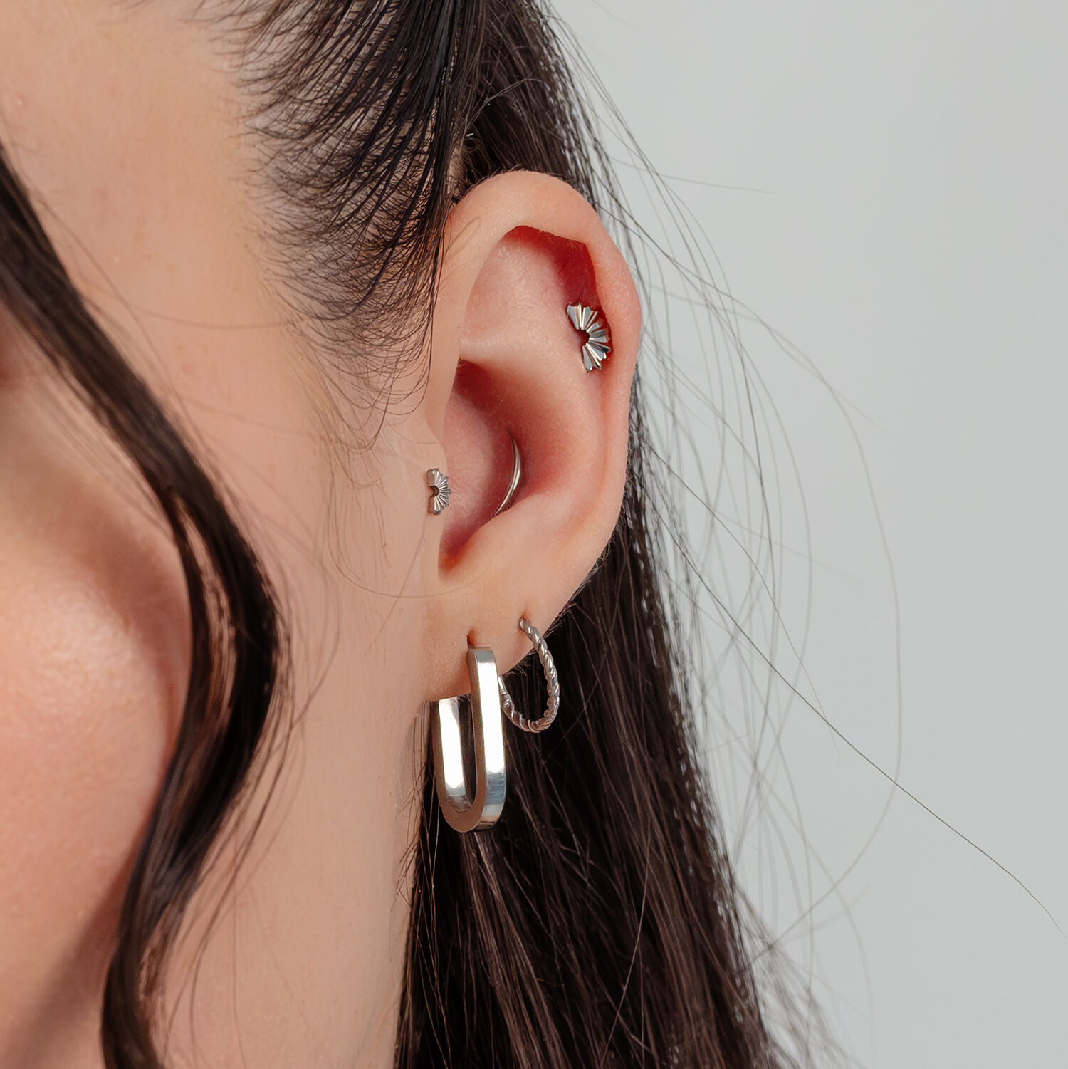 Gros plan d'une oreille avec des boucles d'oreilles argentées et un embout de piercing en forme de rayons de soleil pliés.