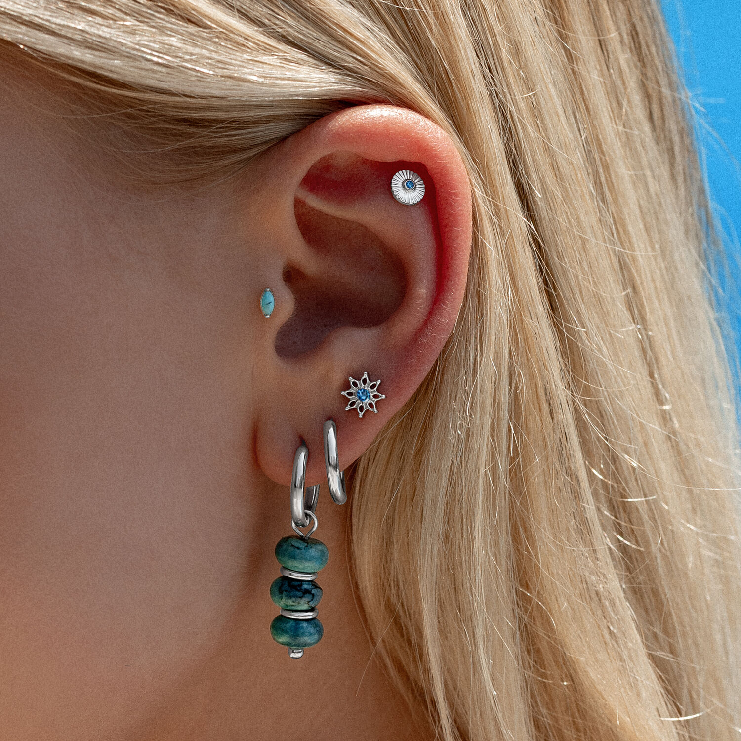 Embout d'oreille en cristal turquoise Oreille avec plusieurs piercings, y compris embout en cristal turquoise et créoles argentées, devant un ciel bleu