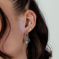 Gros plan d'une oreille avec plusieurs boucles d'oreilles en argent, dont une grande avec opale, sur fond blanc.
