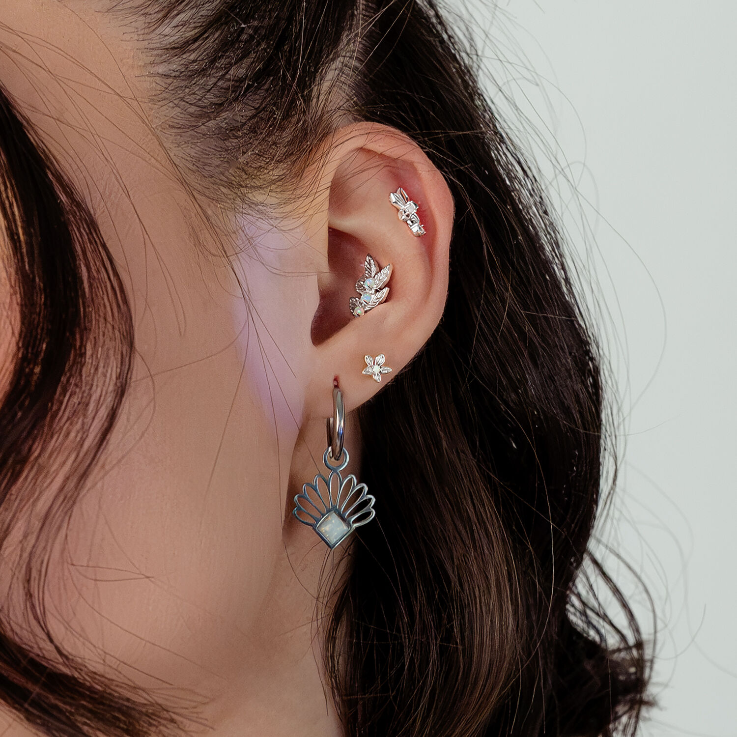 Gros plan d'une oreille avec plusieurs boucles d'oreilles en argent, dont une grande avec opale, sur fond blanc.