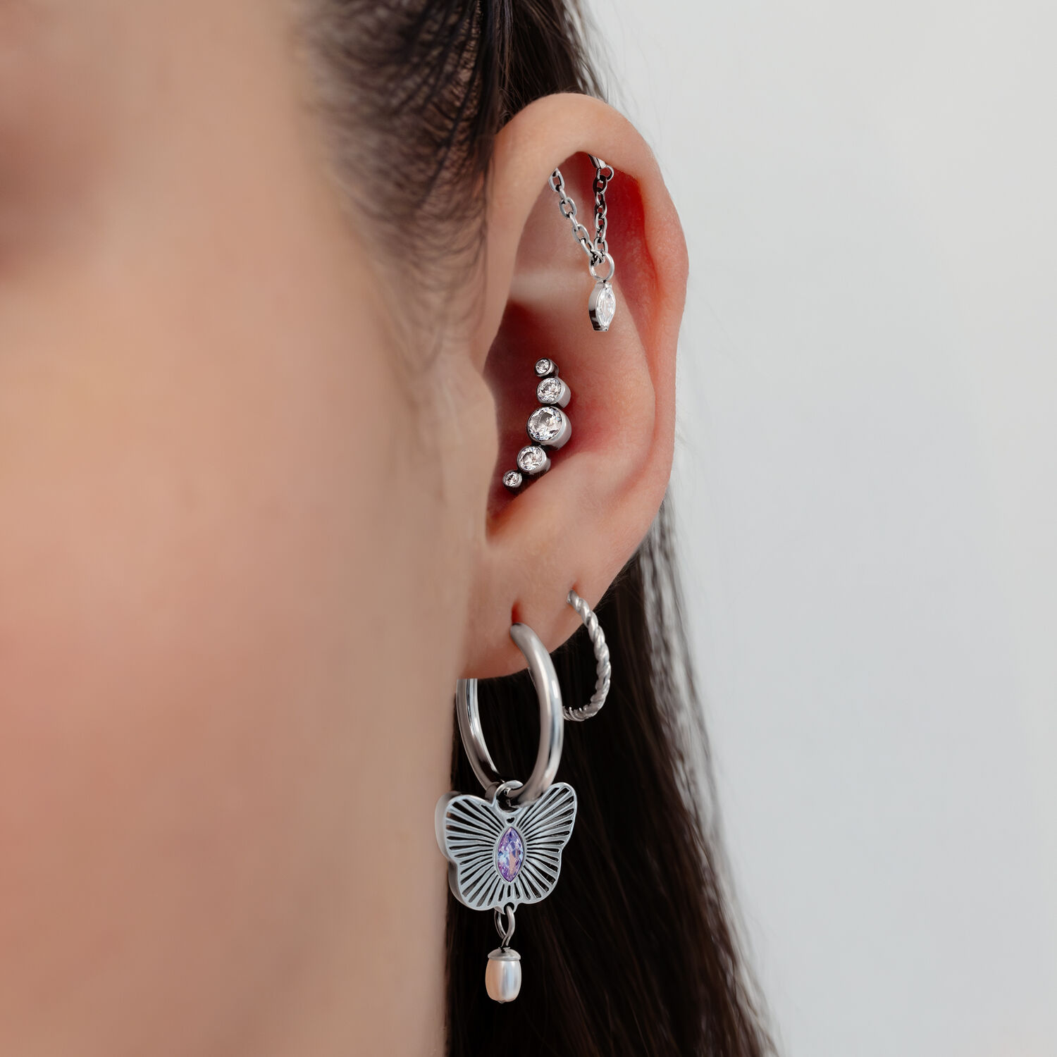 Piercings d'oreille en argent avec cristaux Gros plan d'une oreille avec plusieurs piercings en argent, des cristaux et un pendentif papillon sur fond blanc.